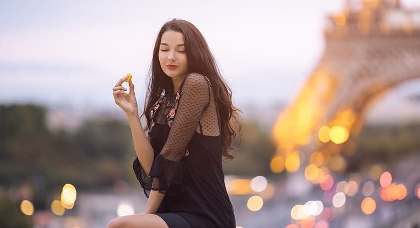 Woman seated outdoors holding a small flower, with the Eiffel Tower blurred in the background at dusk