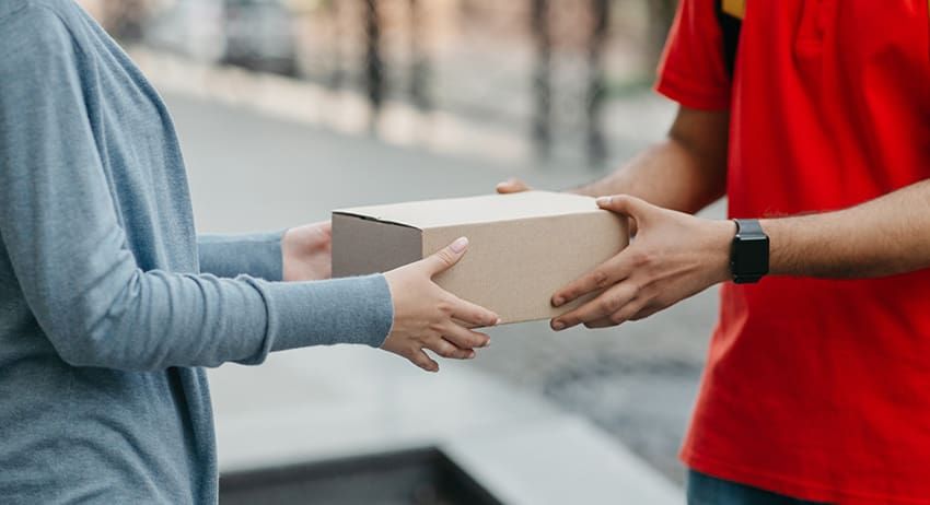 Two people exchanging a small cardboard package outdoors, one in a red shirt and one in a gray sweater