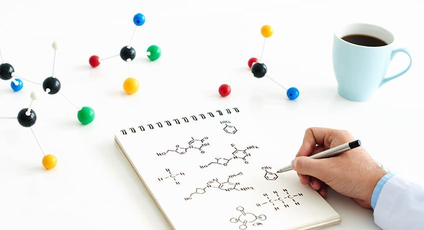 Hand writing chemistry notes in a spiral notebook beside a coffee mug and colorful molecular models