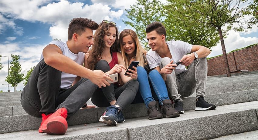 Four friends sitting on outdoor steps, smiling and looking at a smartphone together