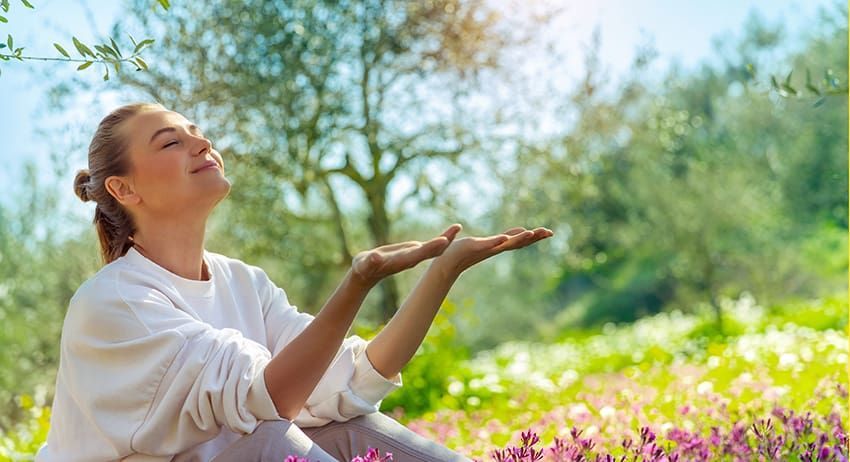Person sitting in a sunlit garden, eyes closed, hands raised in a peaceful pose amid flowers and greenery