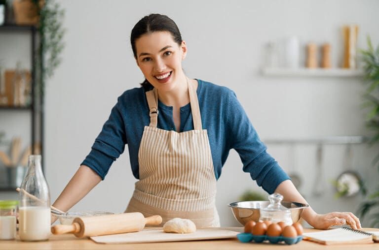 Person in a beige apron smiling while rolling dough in a bright kitchen, with bowls and baking tools nearby