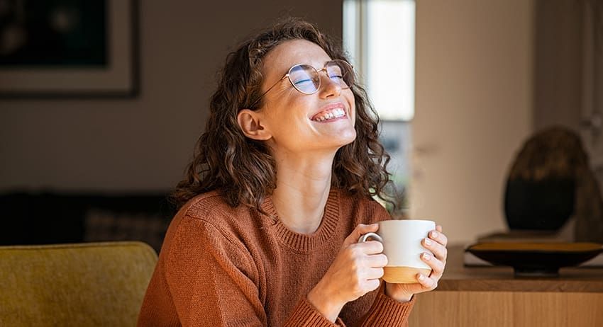 Smiling person in a brown sweater holding a mug indoors by a window