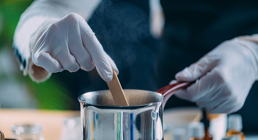 Gloved hands stirring a mixture in a metal beaker with a wooden stick