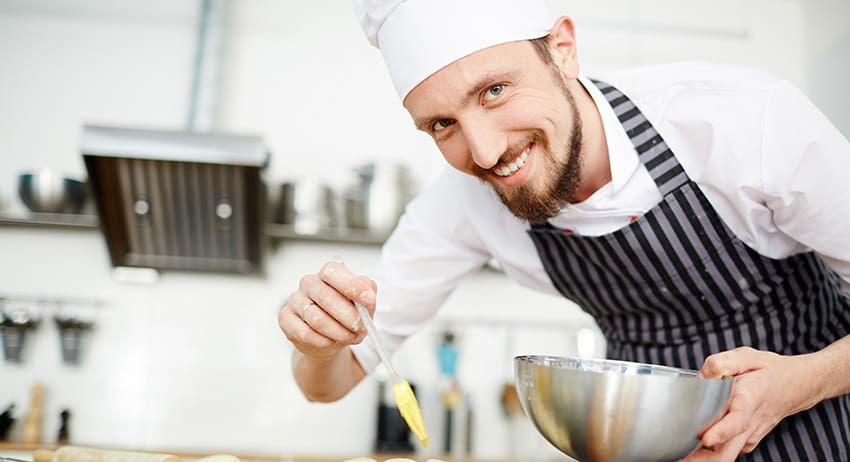 Chef in white uniform and striped apron smiling while mixing ingredients in a kitchen