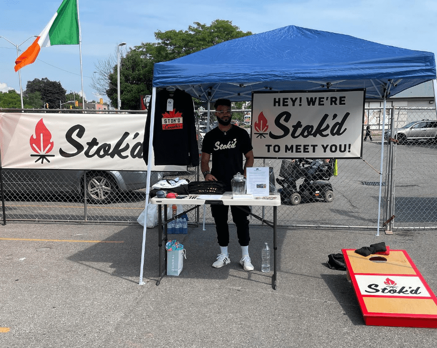 Vendor under blue canopy at Stokd booth with Irish flag, banner, table, and cornhole board.