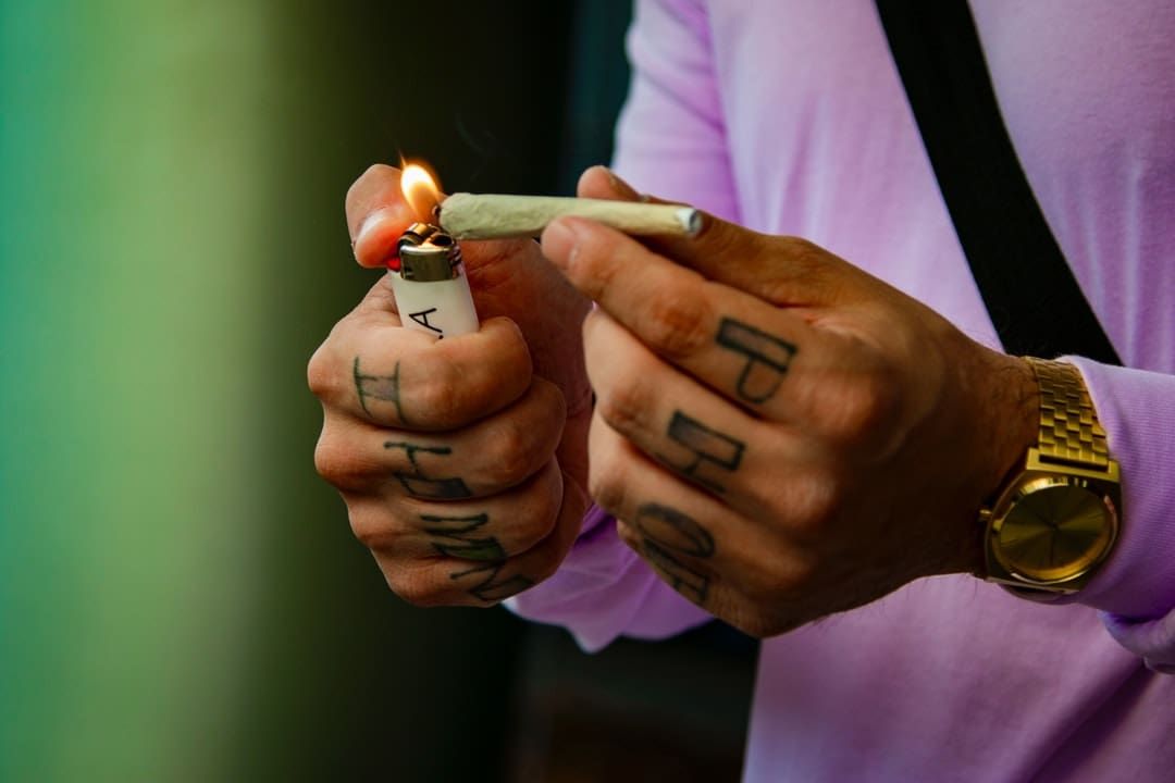 Tattooed hands lighting a cigarette with a lighter, wearing a pink shirt and gold watch