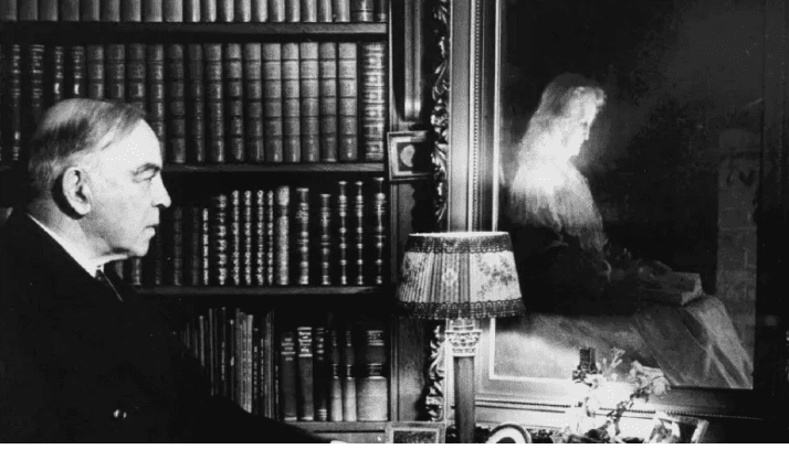 Black-and-white photo of a man in profile beside bookshelves, looking toward a bright, ghostly reflection on glass.