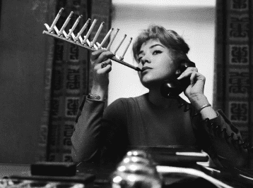 Person holds a comb to their lips in a black-and-white portrait, seated at a table with another comb in hand.