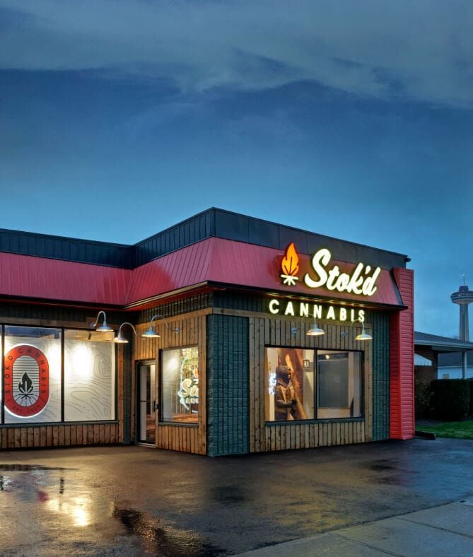 Cannabis dispensary storefront at dusk with red-and-blue facade and lit “Stoki” sign