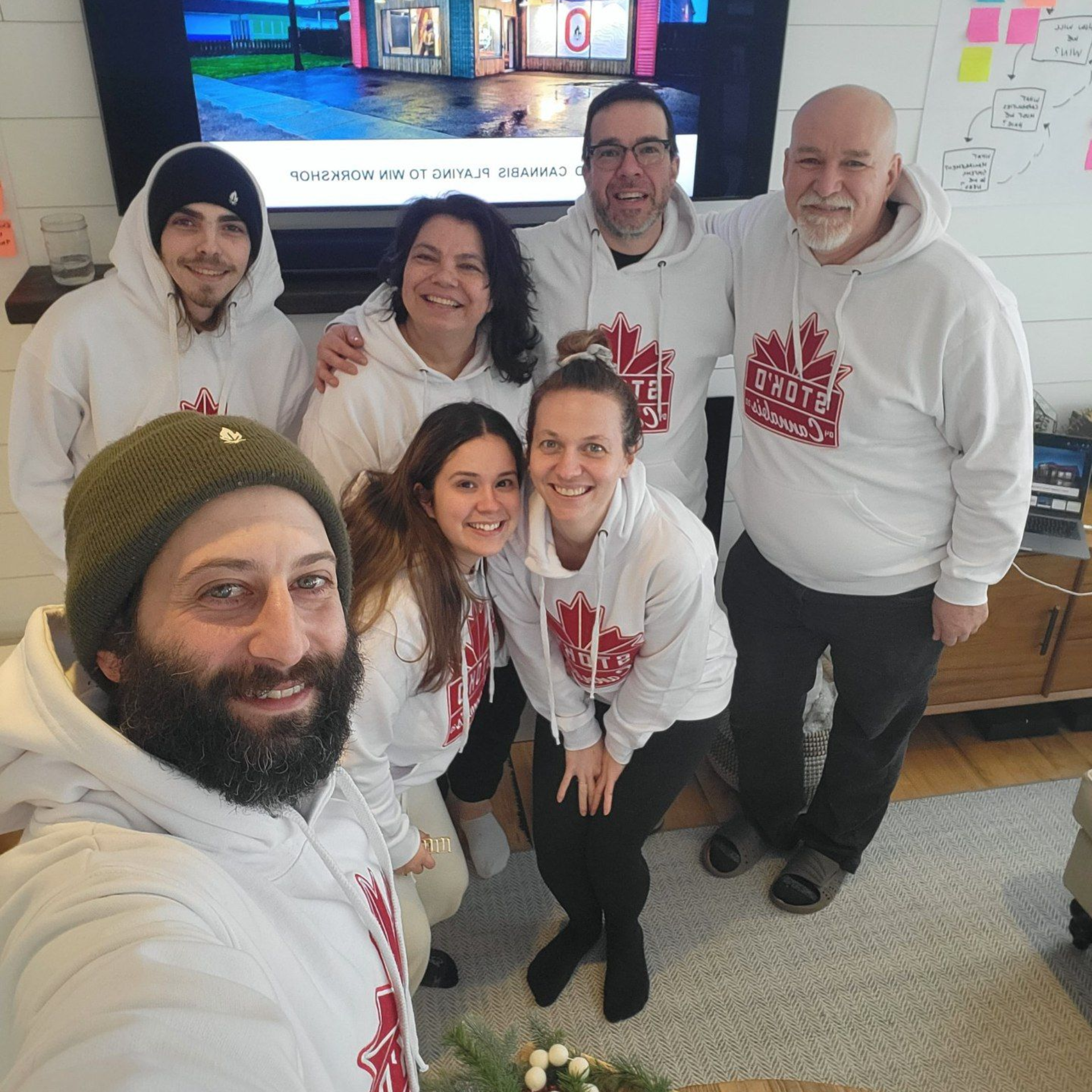 Six people in matching white hoodies pose smiling indoors for a group selfie.