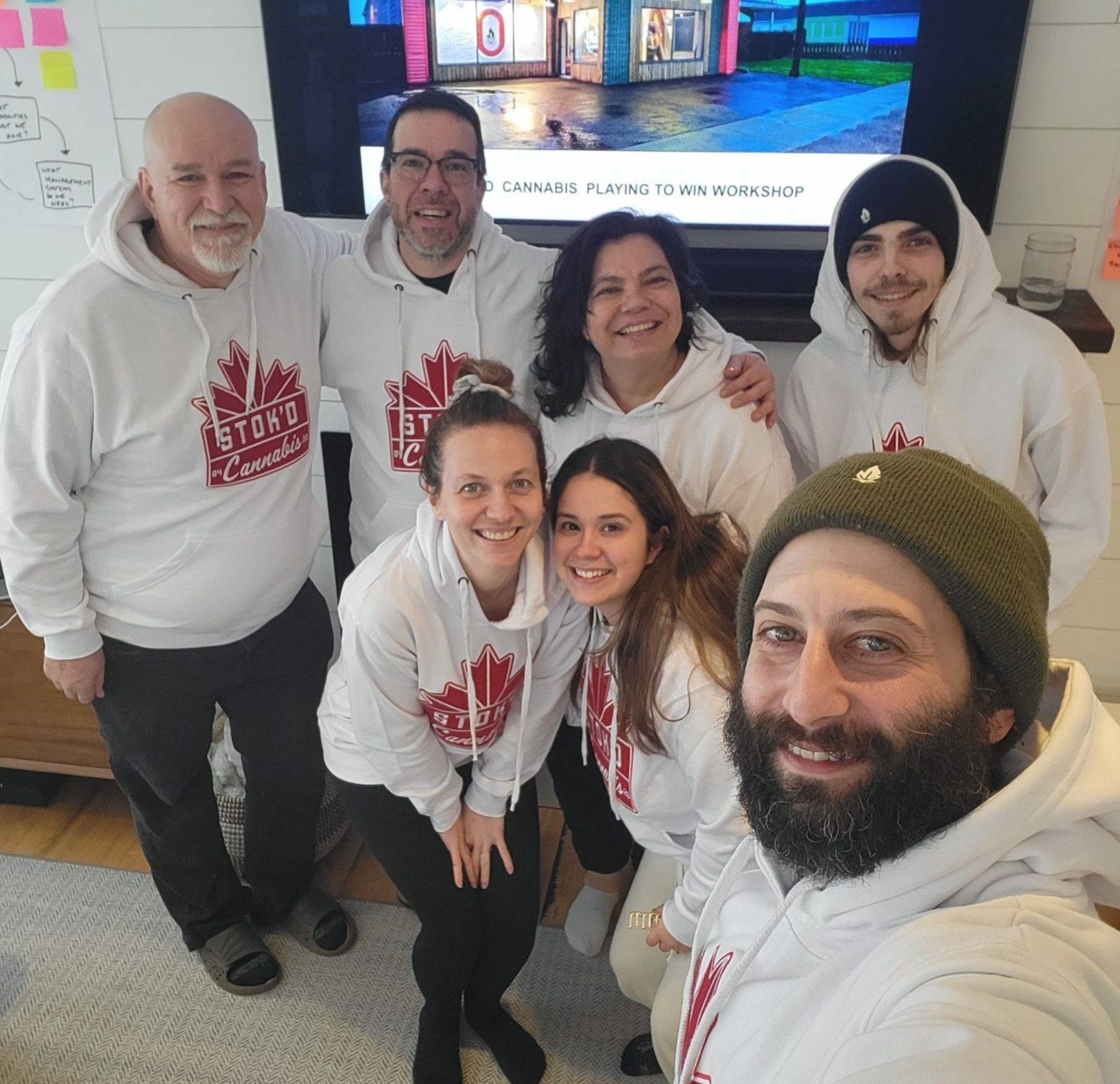 Seven people in white hoodies posing for a selfie indoors, smiling in front of a large screen.