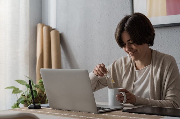 Person stirring a mug beside a laptop at a desk in a bright room