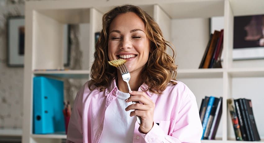 Woman smiling in a pink shirt holding a forkful of pasta in a home kitchen