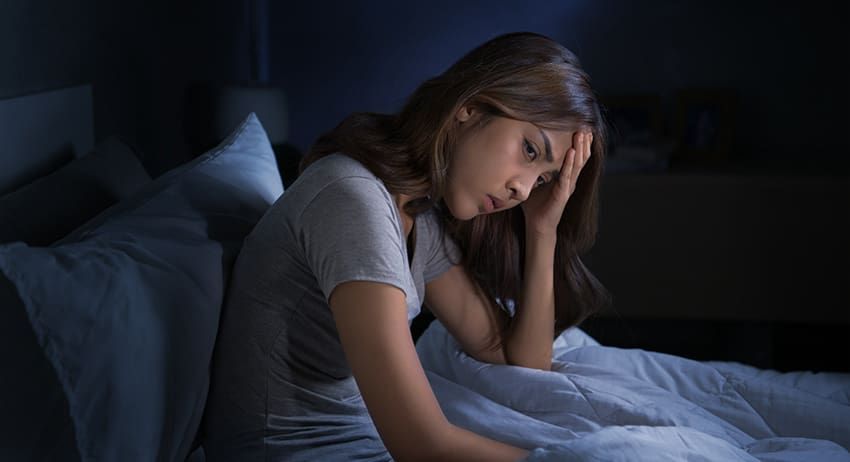 Woman sitting on a bed in dim light, resting her head in her hand and looking upset