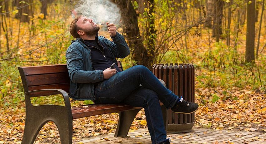 Person sitting on a park bench smoking, with autumn trees and fallen leaves in the background.