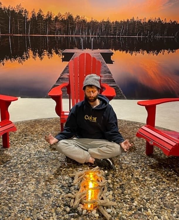 Person sitting cross-legged between red chairs by a reflective lakeside at sunset.
