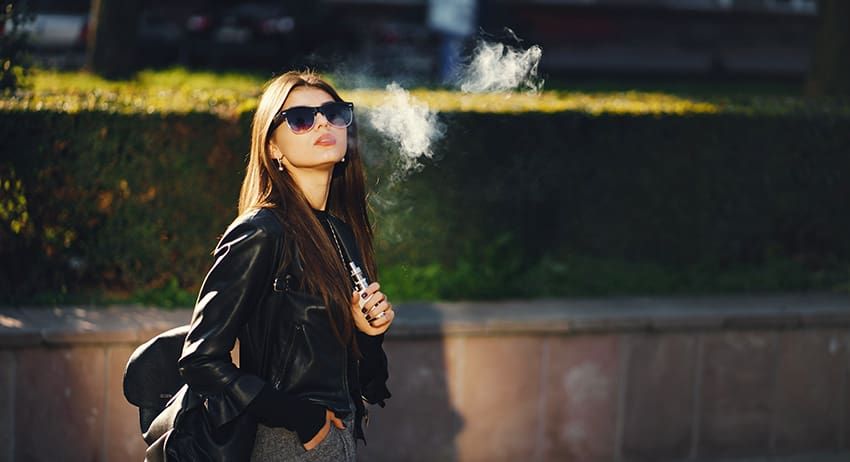 Woman in sunglasses smoking outdoors beside a hedge, wearing a black jacket and backpack.