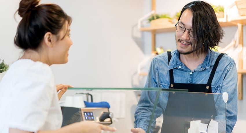 Two people talking across a café counter, with one holding a cup and the other smiling behind a laptop