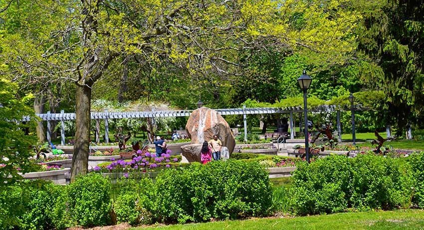 Sunny park with green trees, flower beds, benches, and people near a central fountain