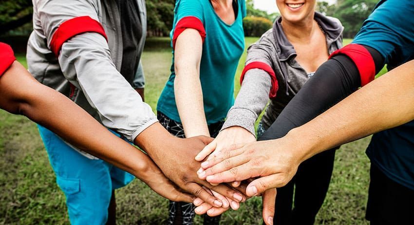 Team members stacking hands together outdoors on grass, smiling in colorful athletic shirts