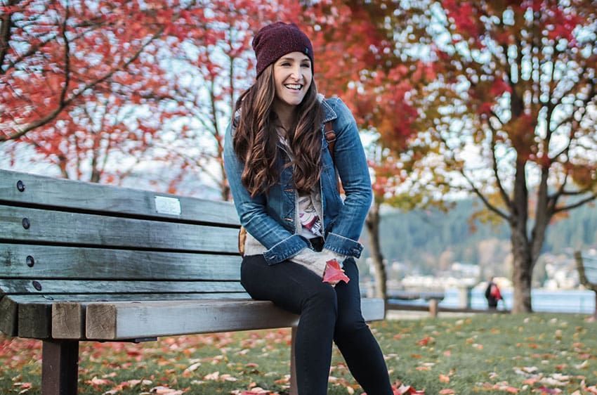 Smiling woman in a maroon beanie sits on a park bench with autumn trees in the background