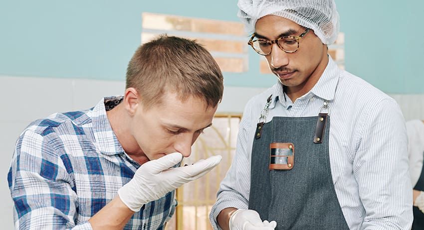 Technician smelling liquid in lab while colleague observes, wearing gloves and protective gear