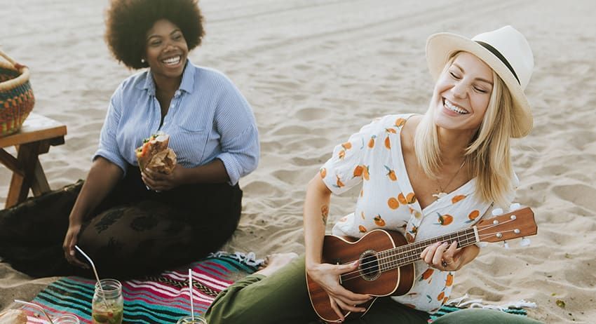 Two people sit on a beach blanket, smiling and playing a ukulele by the water.