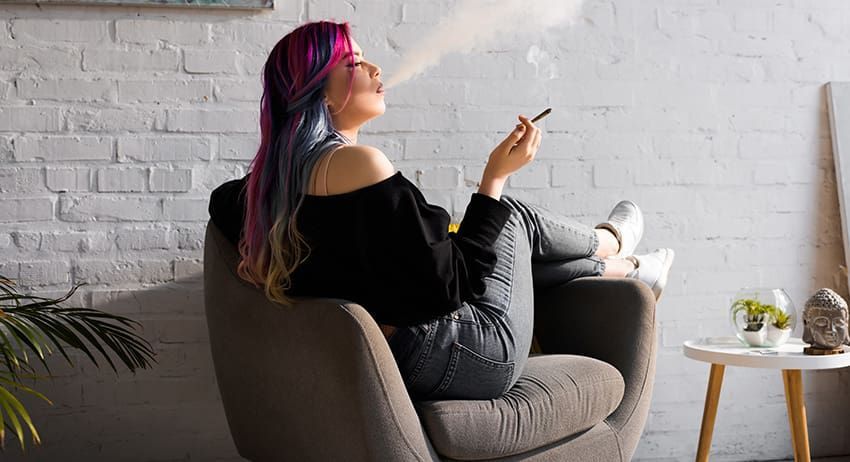 Person with pink hair sitting in a chair, smoking and looking at a phone in a cozy room.