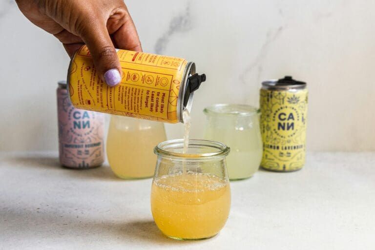 Hand pouring yellow beverage from can into glass beside other cans and glasses