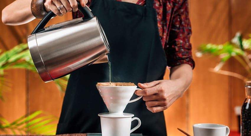 Barista pours hot water from a kettle through a filter into a white cup on a counter