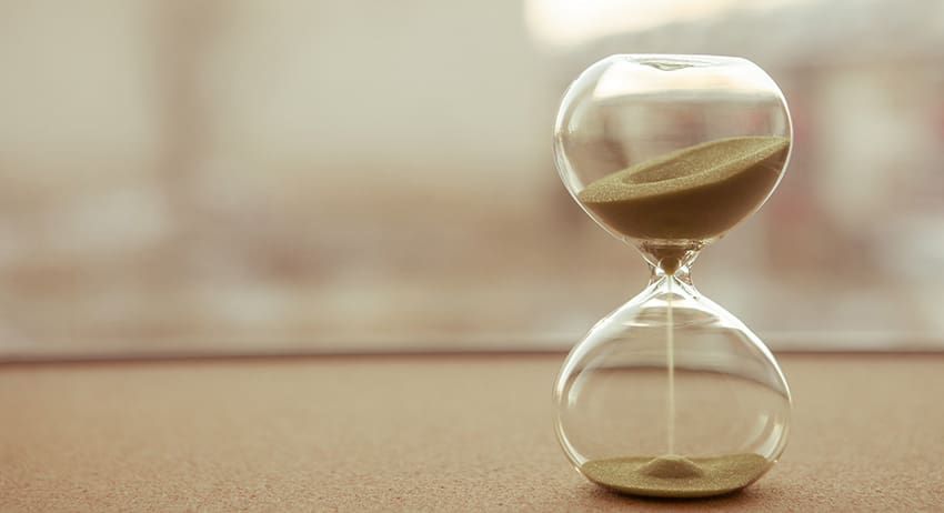 A glass hourglass with sand, standing on a surface against a blurred background.