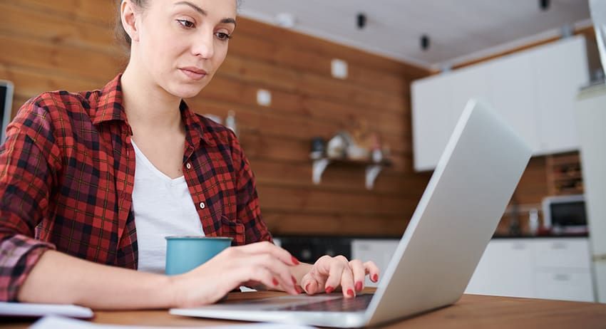 Person typing on a laptop at a wooden table in a kitchen, wearing a red plaid shirt.