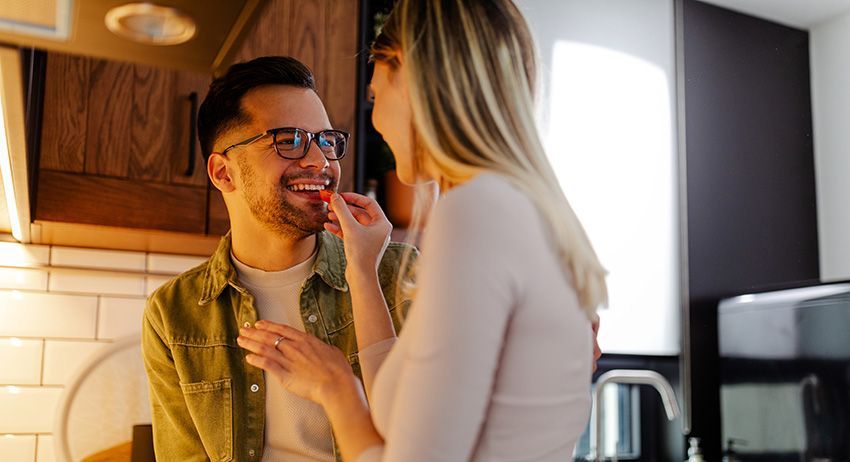 Two people smiling in a kitchen, one feeding the other a bite of food