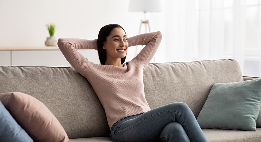 Woman relaxing on a sofa in a bright living room, leaning back with her hands behind her head.