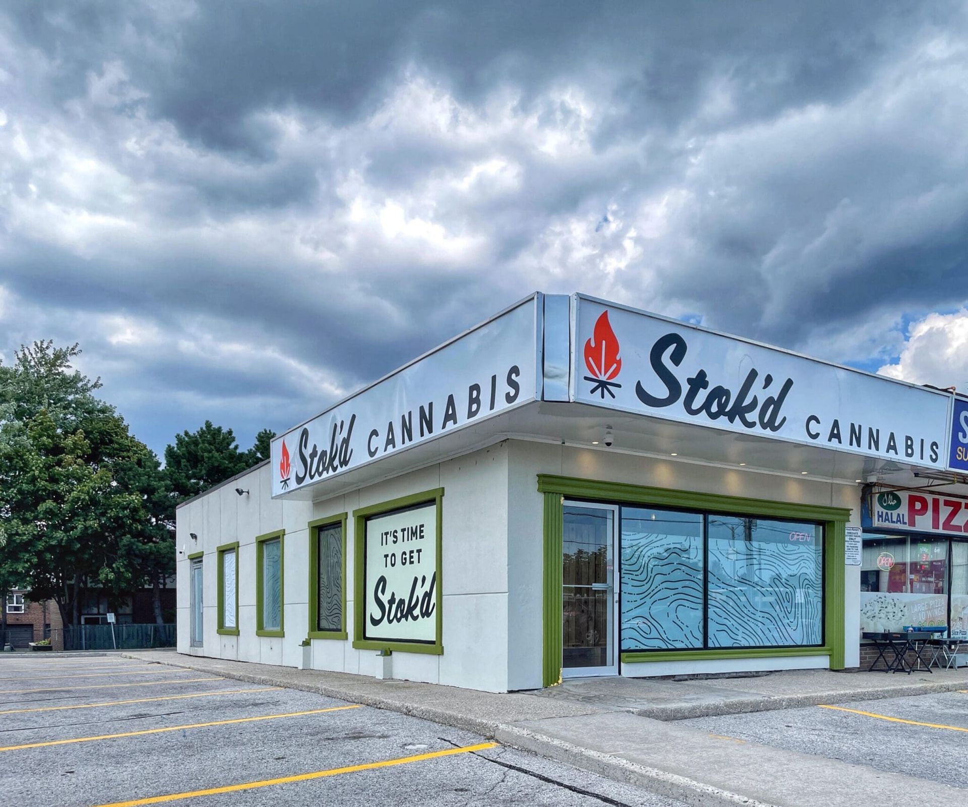 Corner cannabis shop with white exterior and green trim under a cloudy sky, signs reading “Stoked Cannabis.”