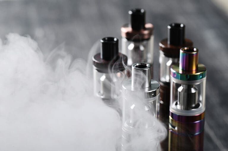 Four colorful vape tanks releasing white vapor on a dark background