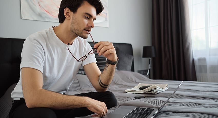 Man sitting on a bed, holding headphones and looking at a laptop in a bright bedroom