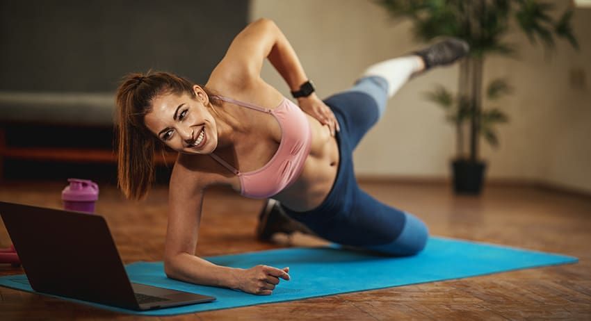 Woman doing side plank on yoga mat beside laptop in a home workout setting