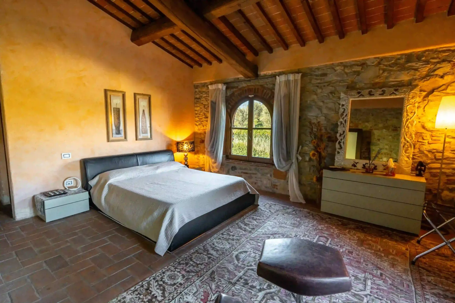 Una camera da letto in stile rustico presenta un letto con testiera nera, pareti in pietra a vista, travi in legno a vista sul soffitto e pavimento piastrellato.