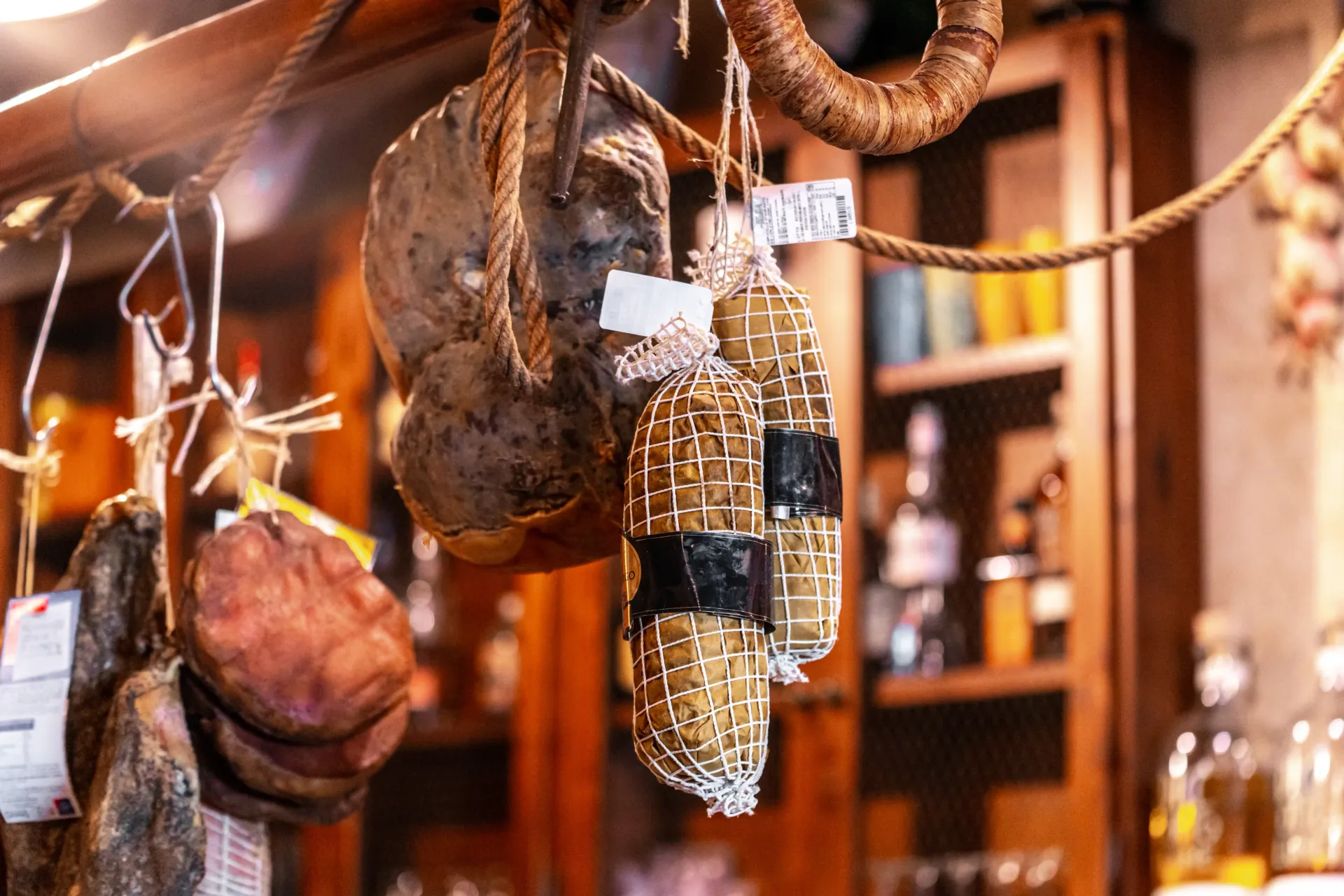 In un negozio rustico e scarsamente illuminato, con scaffali di legno sullo sfondo, vari tipi di salumi sono appesi a delle corde.