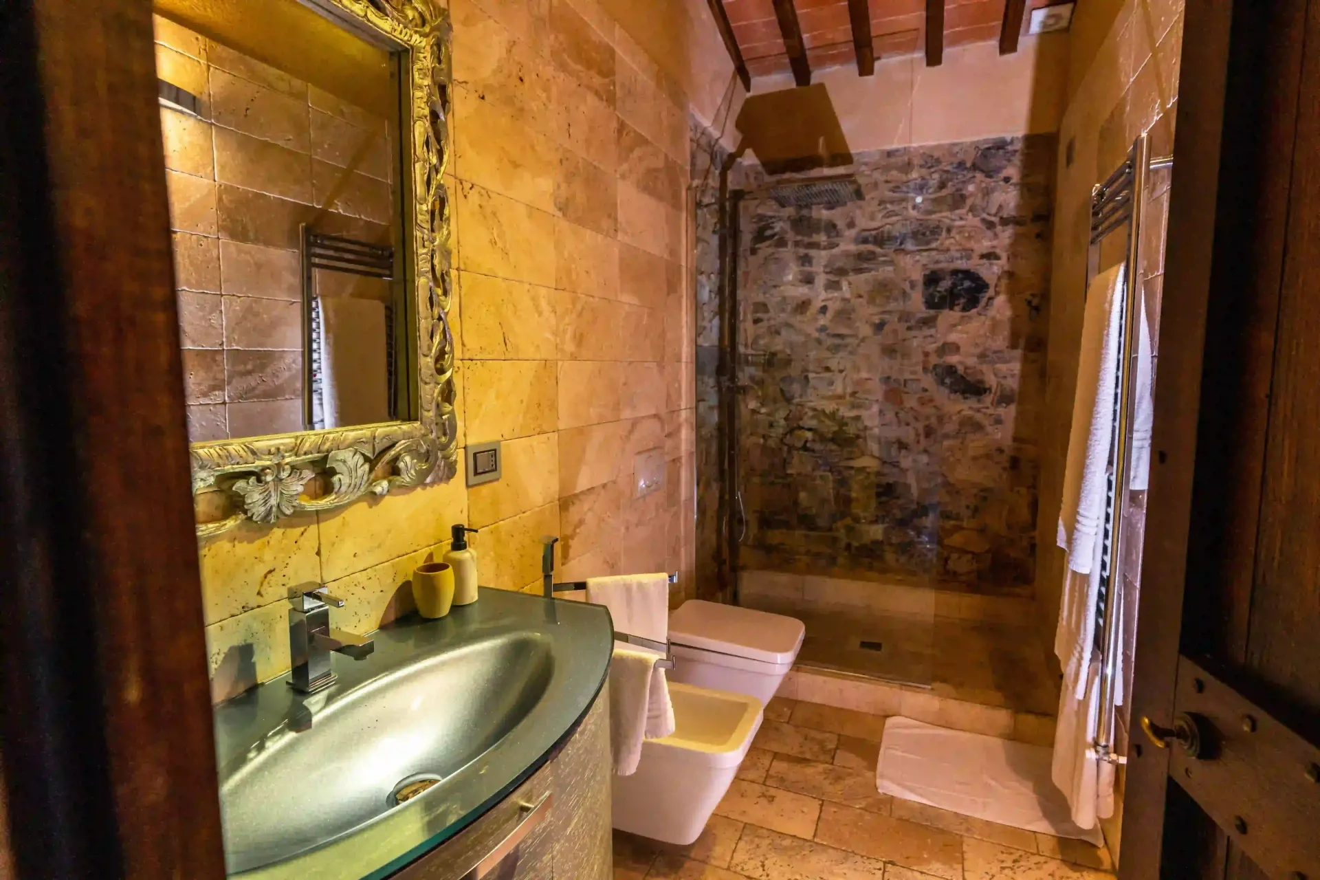 Un bagno in stile rustico con pareti in pietra color sabbia, un lavabo a ciotola in argento, uno specchio con cornice decorata e una cabina doccia.