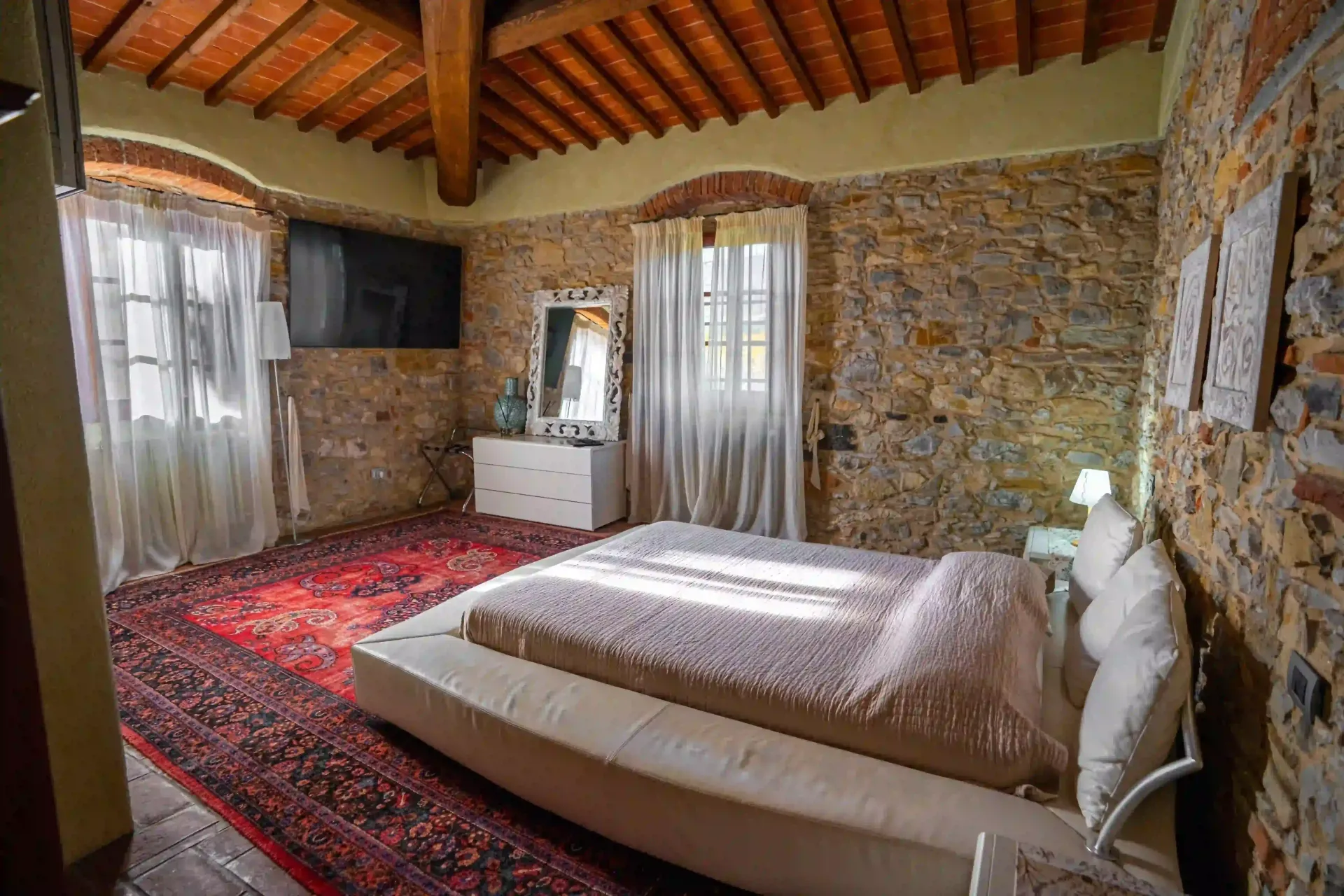 Una camera da letto con pareti in pietra rustica, travi a vista sul soffitto, un grande tappeto rosso e un letto basso con lenzuola bianche.