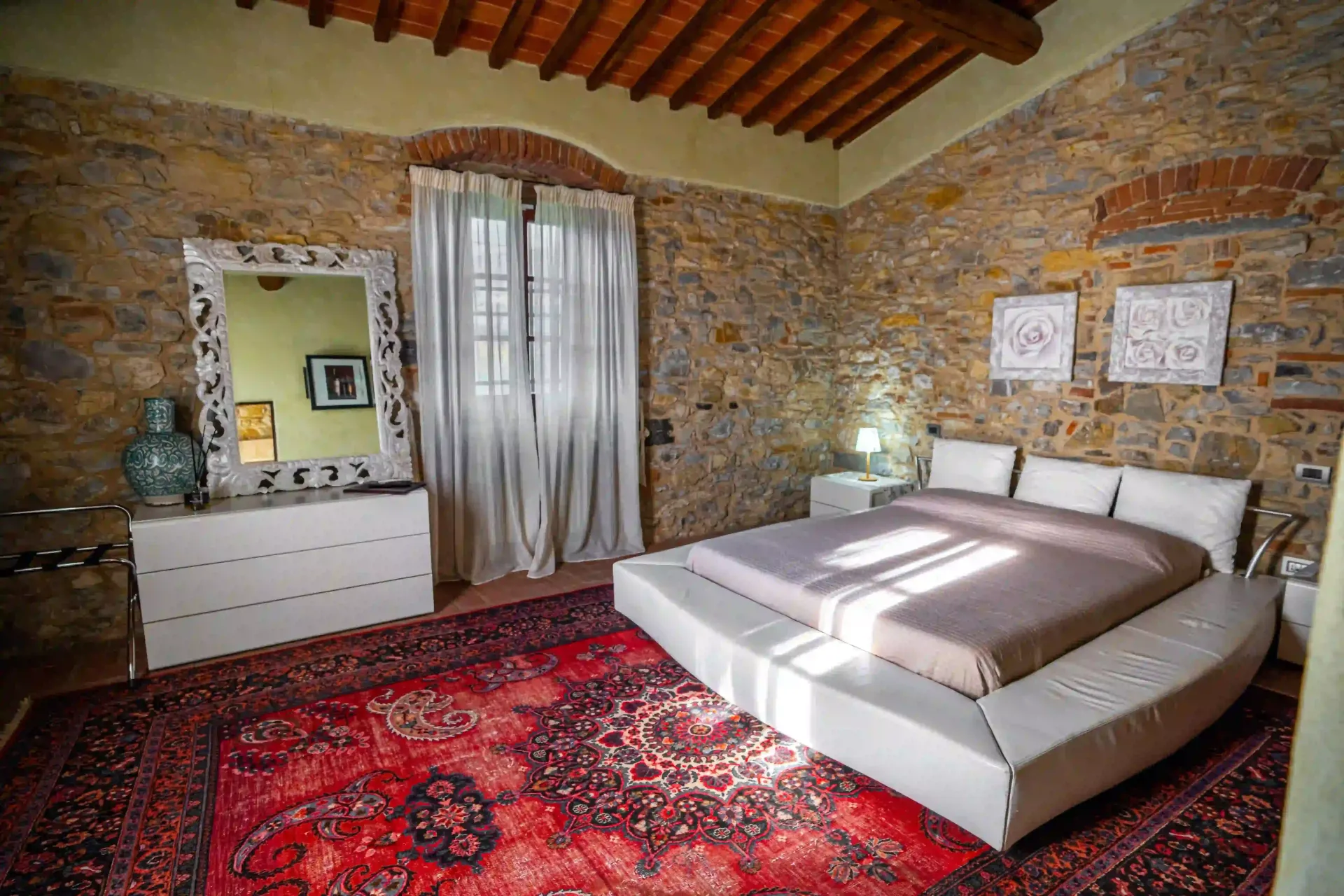 Una camera da letto in stile rustico presenta pareti in pietra, un soffitto in legno, un grande tappeto rosso, un letto a piattaforma e una cassettiera con specchio.