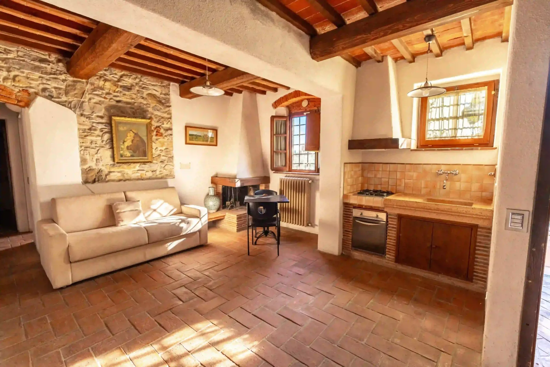 Un soggiorno rustico con divano beige, parete in pietra, pavimento in cotto, cucina a vista e soffitto con travi in legno.