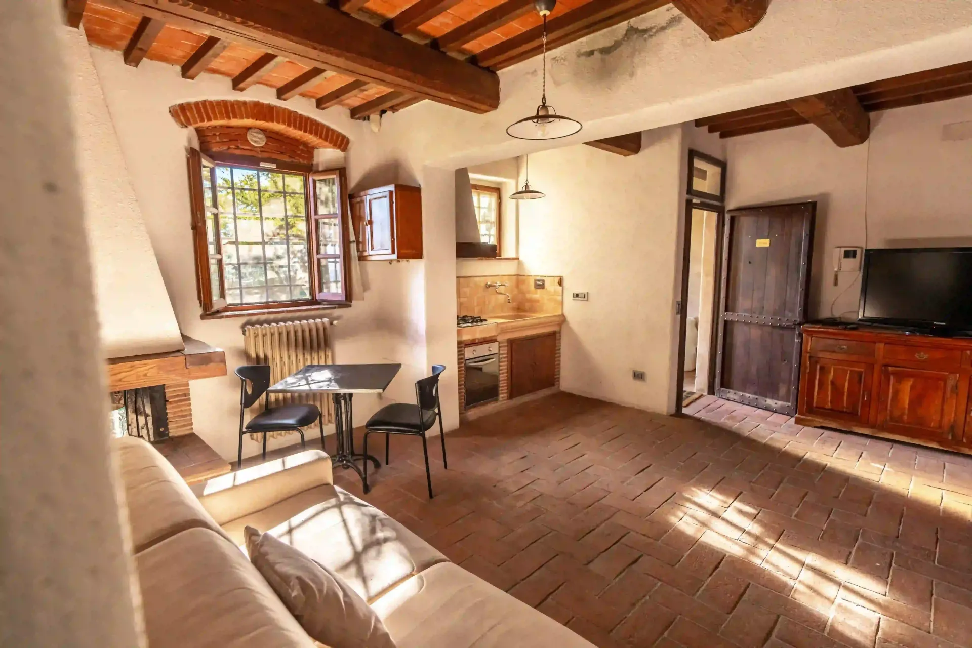 Un soggiorno rustico con pavimento piastrellato, travi in legno a vista, un piccolo tavolo da pranzo, un angolo cottura e un divano.
