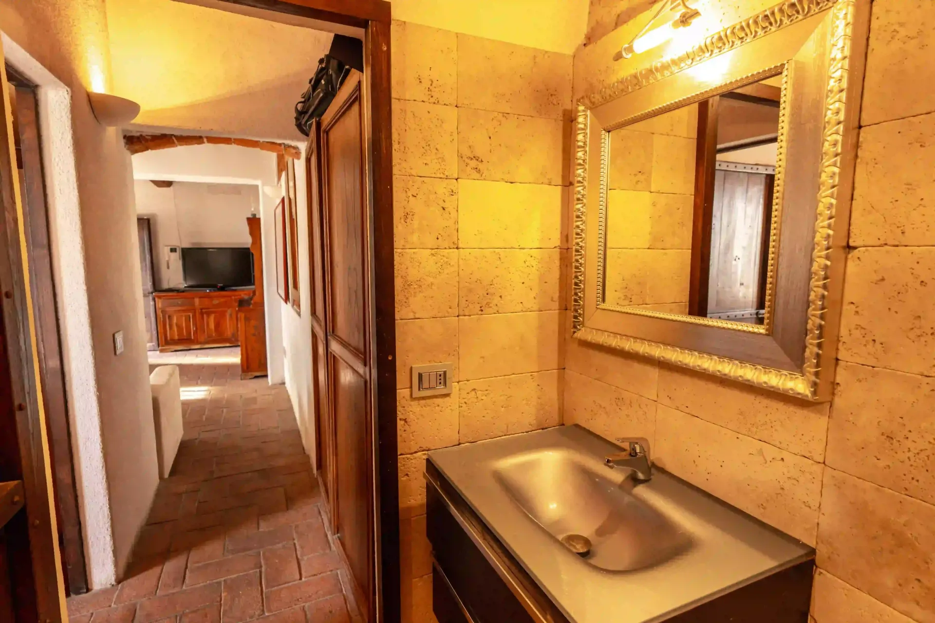 Un mobile da bagno con specchio con cornice dorata e parete rivestita in pietra, che si apre su un corridoio con vista sulla zona giorno.