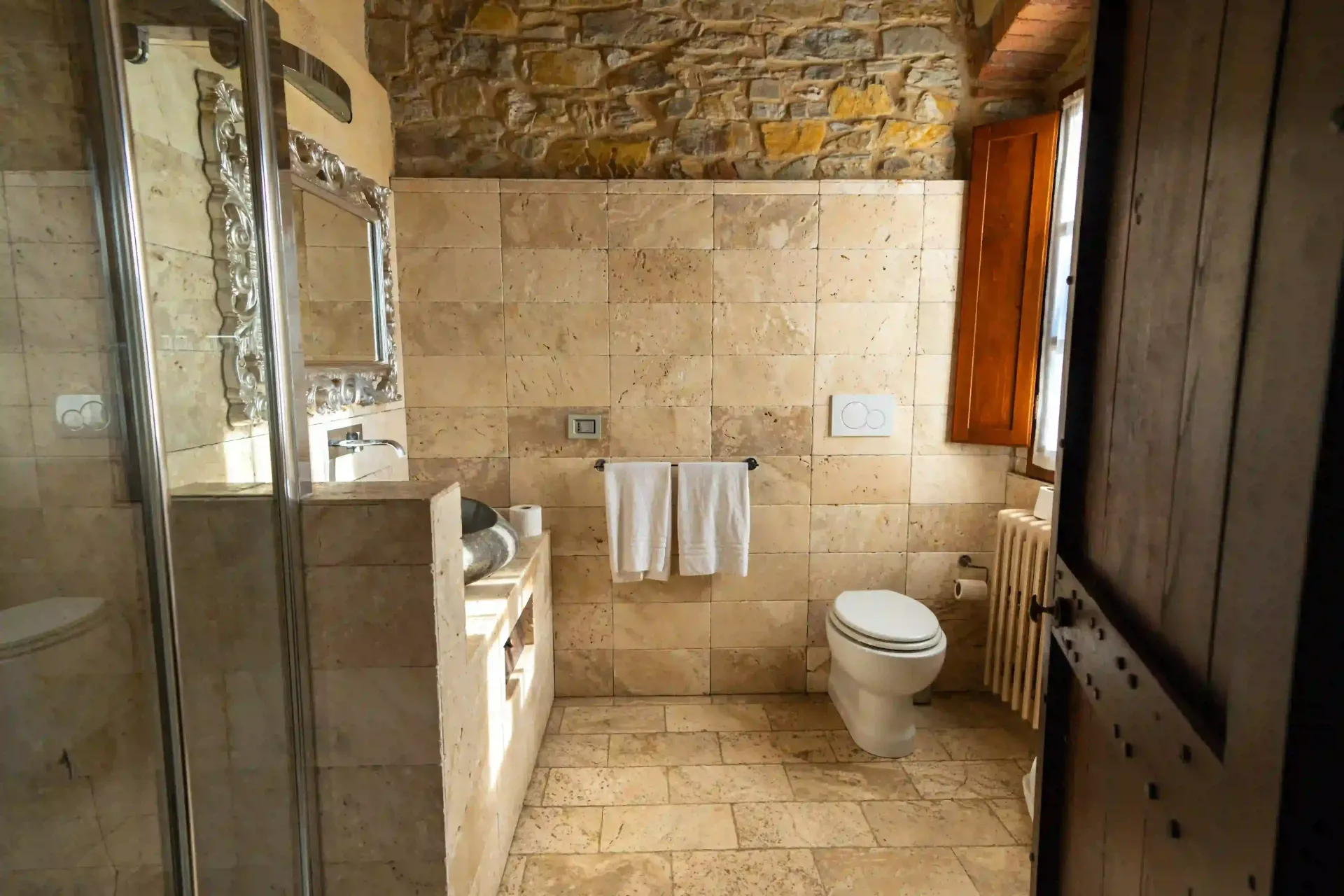 Un bagno rustico caratterizzato da pareti in pietra chiara e pavimento piastrellato, una cabina doccia in vetro, un mobile lavabo e un WC bianco.
