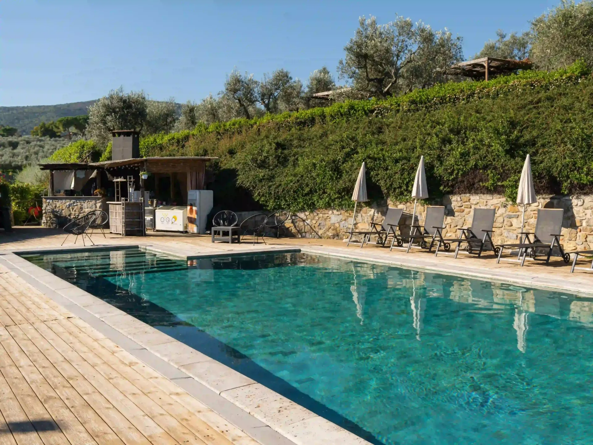 Una piscina rettangolare con acqua cristallina color turchese, circondata da una terrazza in legno, lettini prendisole e con lo sfondo di una collina.