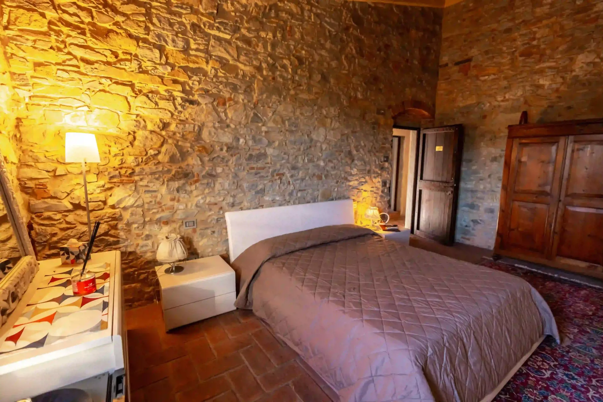 Una camera da letto rustica con pareti in pietra, un letto matrimoniale con una trapunta grigia, un comodino e un armadio in legno scuro.